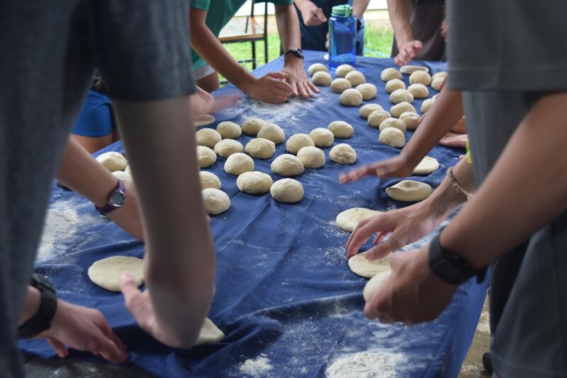 A group of people are gathered around a blue-covered table, kneading and shaping dough into small, round balls. Their hands are actively working on the dough, which is scattered across the table. The scene suggests a collaborative cooking activity, possibly preparing food together. Some individuals are wearing watches, and the focus is on the hands and the dough.